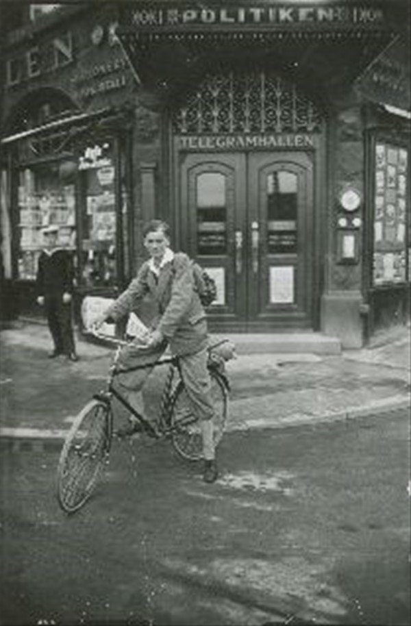 Motiv: Foran indgangen ses en ung mand på en tilsyneladende ny cykel med vimpel